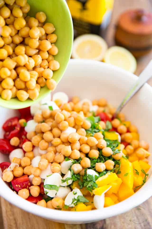 Chickpeas getting added to bowl containing chopped raw veggies and couscous